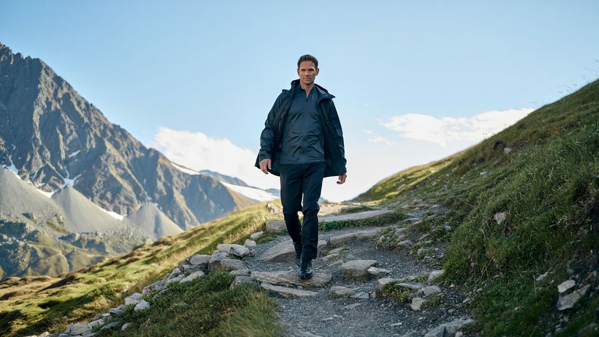 Randonneuse adulte portant une veste coupe vent unie sur sentier de montagne sans logo ni texte